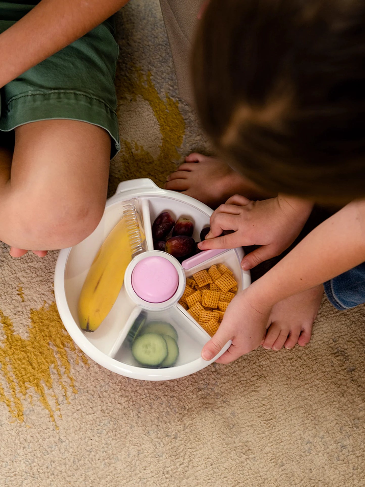 Children using the GoBe Snack Spinner with fruits, cereal, and a cucumber on a cozy carpet.