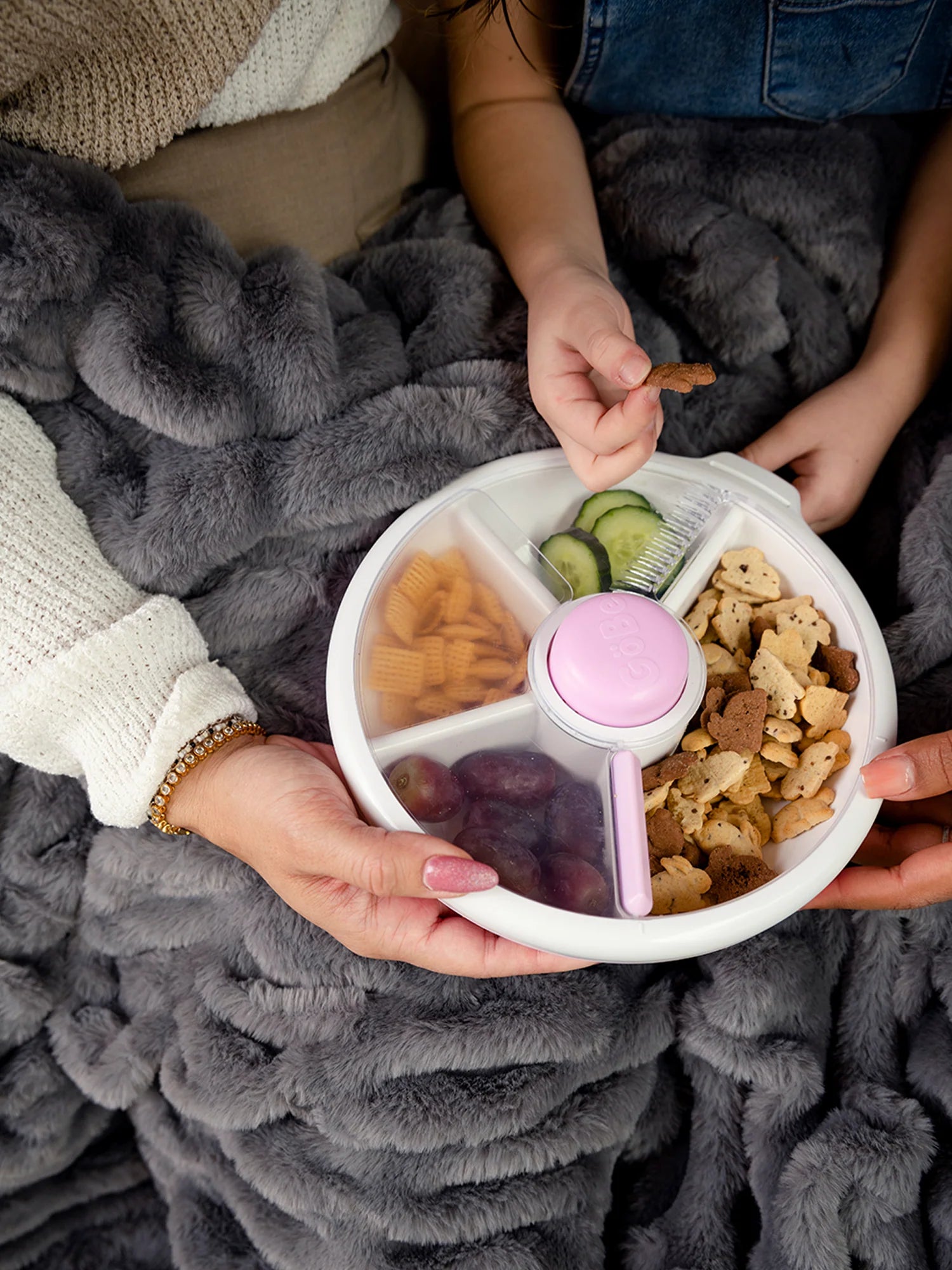 GoBe Snack Spinner in lilac with assorted snacks, held by hands under a cozy blanket.