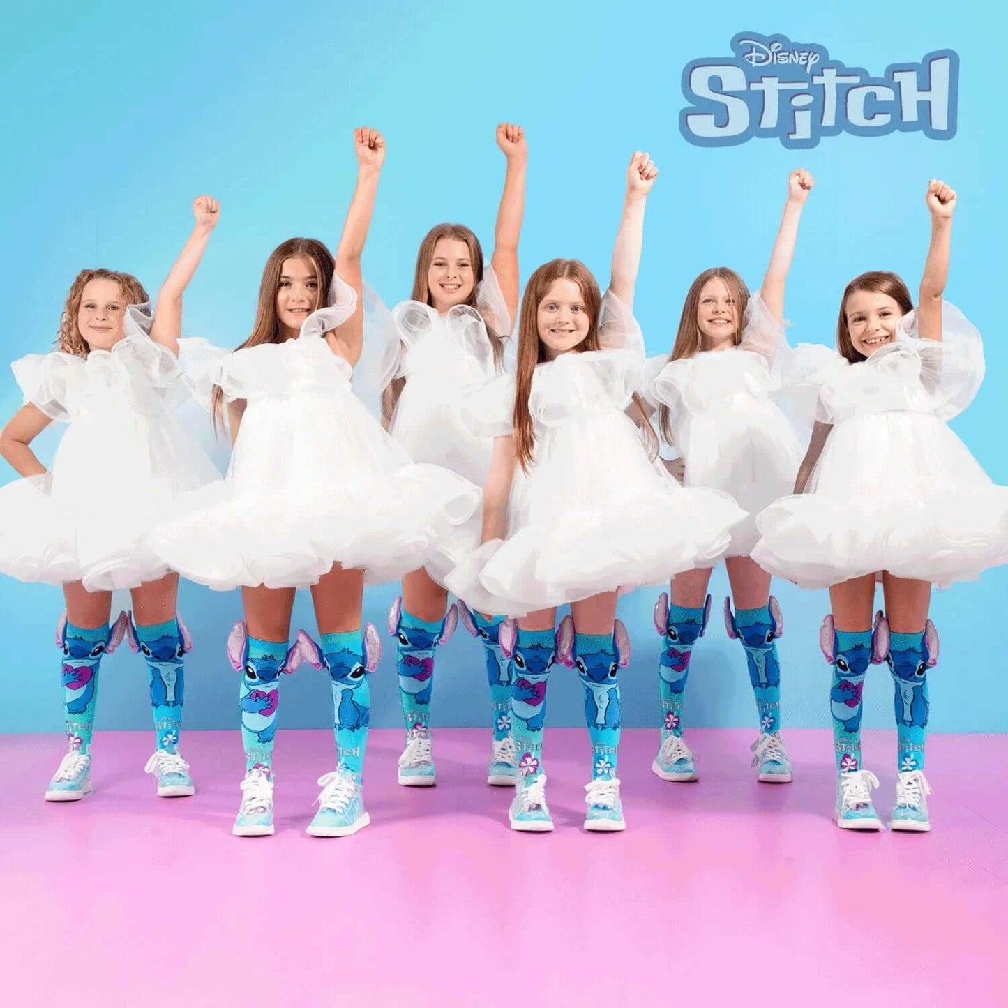 Grupė mergaičių baltomis suknelėmis demonstruoja spalvingas Disnėjaus Stičo iki kelių siekiančias kojines, švęsdamos iškeltomis rankomis.